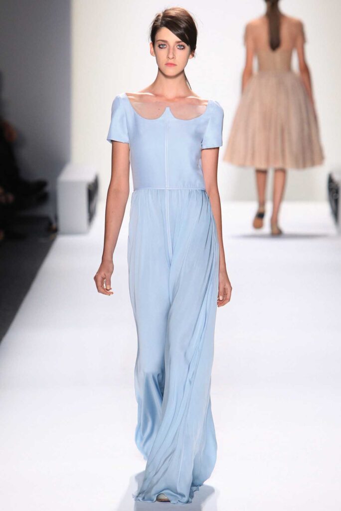 Woman in a light blue gown walks on a fashion runway; another model is visible in the background.