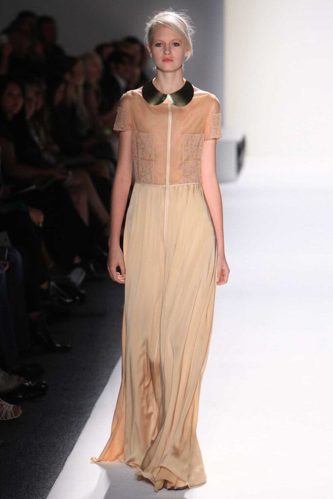Model walking runway wearing a beige gown with sheer sleeves and a dark, rounded collar.