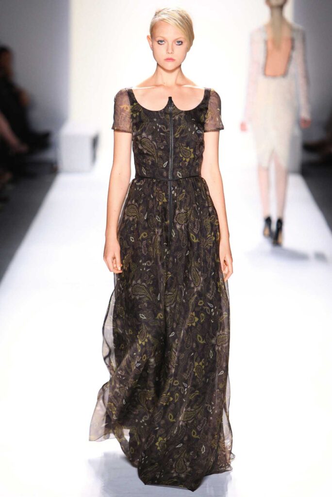 Model in sheer, patterned black gown with short sleeves walking on a white runway during a fashion show.