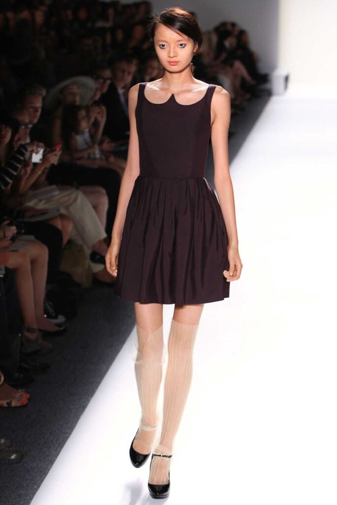 Model in a black sleeveless dress and sheer knee-high socks walks down a white runway at a fashion show.