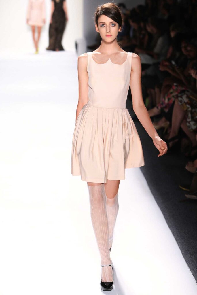 Model in a light pink sleeveless dress with knee-high socks and black heels walks on a fashion runway.