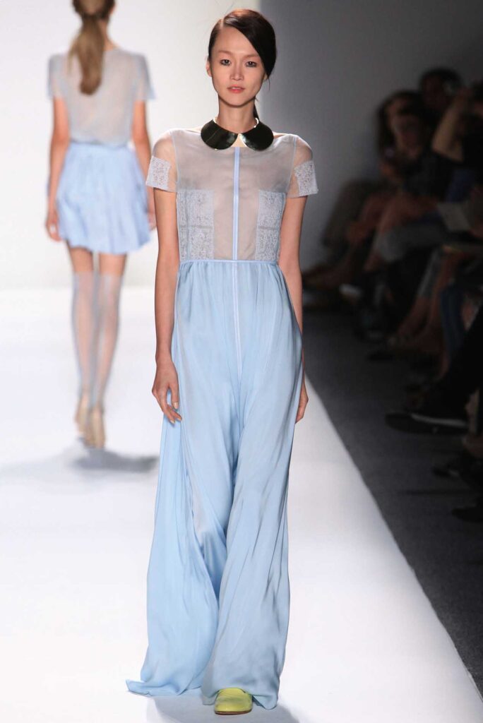 Model in a pale blue sheer dress with a black collar walks down a runway; audience is in the background.