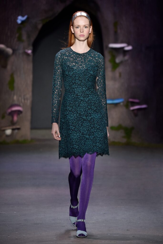 Model in green lace dress, purple tights, and headband walking runway with a dark, whimsical forest backdrop.