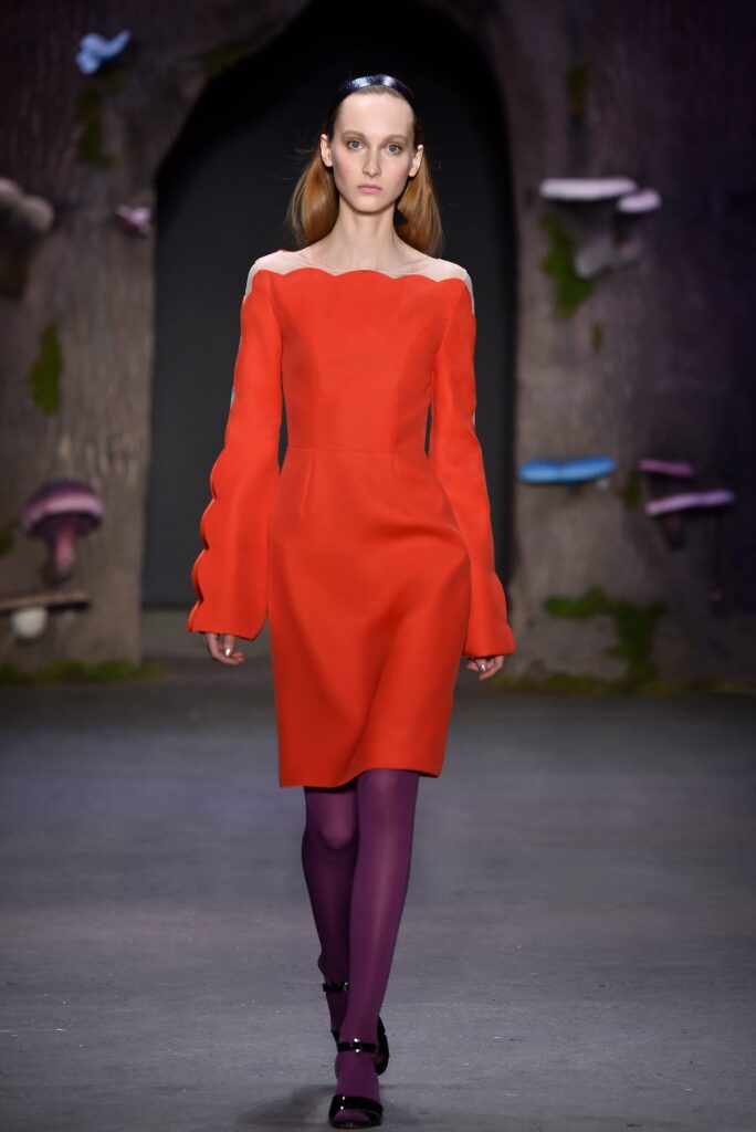 Model in a red dress and purple tights walking a runway with a whimsical, forest-like background.