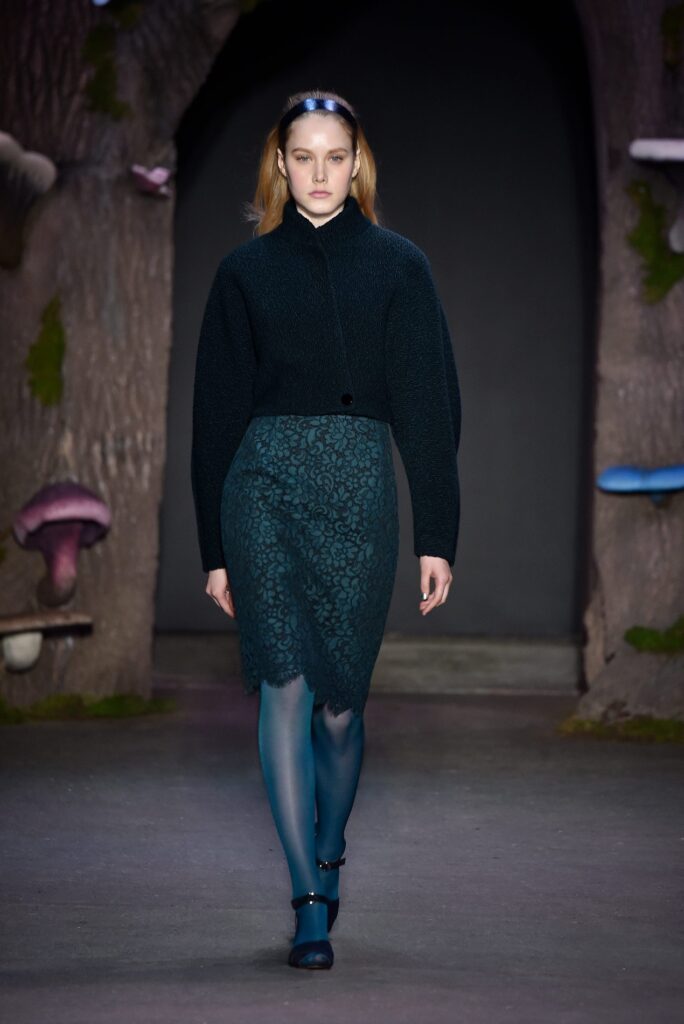 Model walks runway in dark sweater, teal lace skirt, matching tights, and headband; forest set with mushrooms.