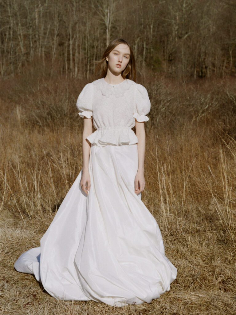 Woman in a white dress stands in a dry, grassy field with trees in the background.
