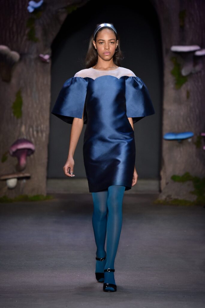 Model in a navy blue dress with puffy sleeves, teal tights, and headband walks on a whimsical runway.