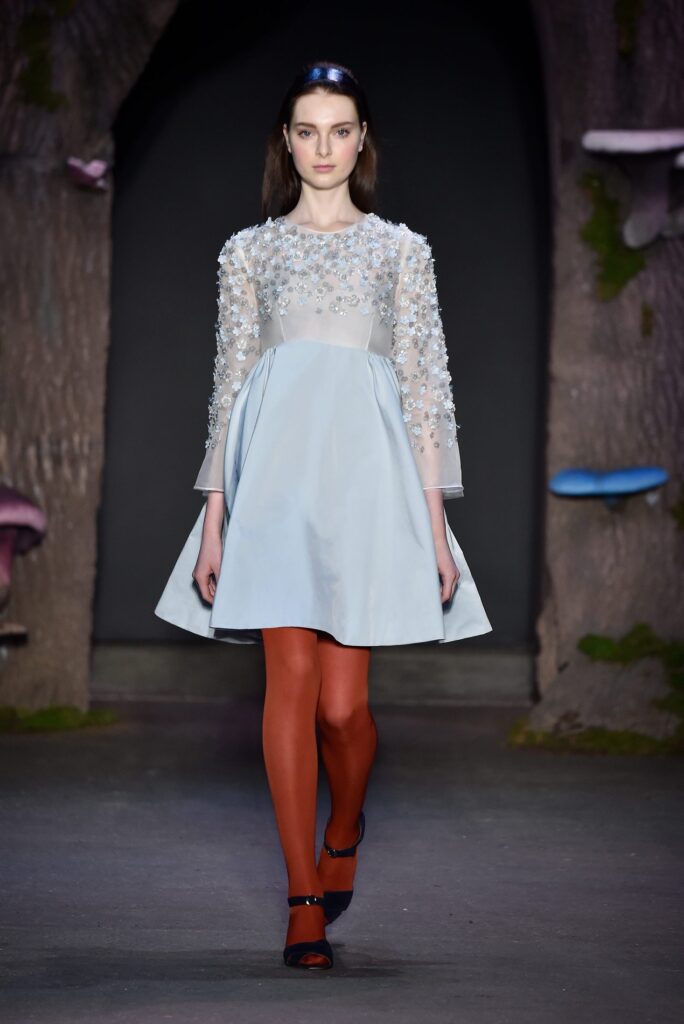 Woman on runway wears a light blue dress with floral details, orange tights, and black heels.