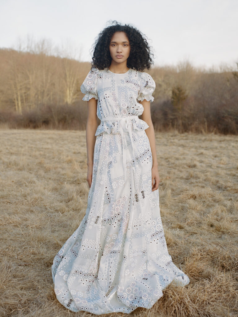 Woman in a white, floral-patterned dress stands in a dry grassy field with trees in the background.