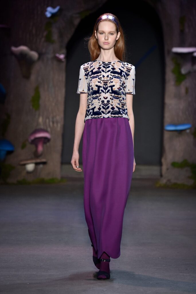 Model in a floor-length purple skirt and patterned top walks a forest-themed runway with large mushrooms.