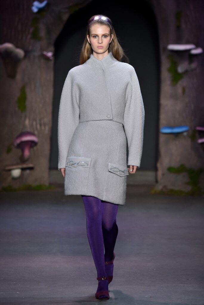 Model in a gray textured coat with purple tights walks on a runway with a fantasy forest backdrop.
