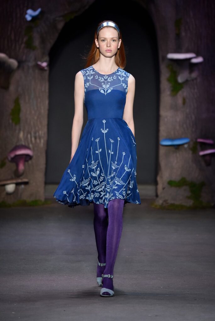 Model in a blue dress with white floral patterns, purple tights, and headband walks on a fantasy-themed runway.