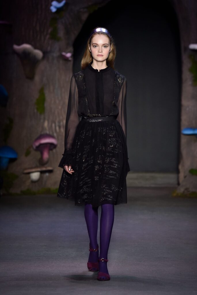 Model in a black lace dress, purple tights, and burgundy shoes walks on a runway with a forest backdrop.