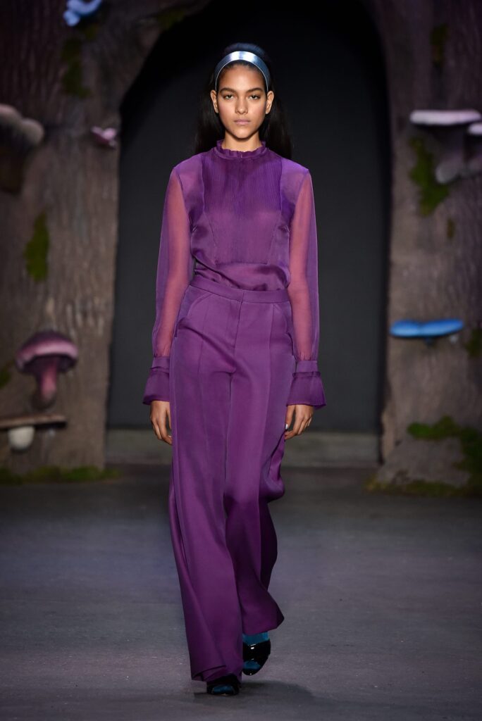 Model walks runway in sheer purple blouse and matching wide-leg pants, headband, woodland backdrop with mushrooms.