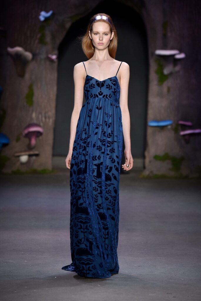 A woman in a long, dark blue patterned dress walks on a runway with a forest-themed backdrop.