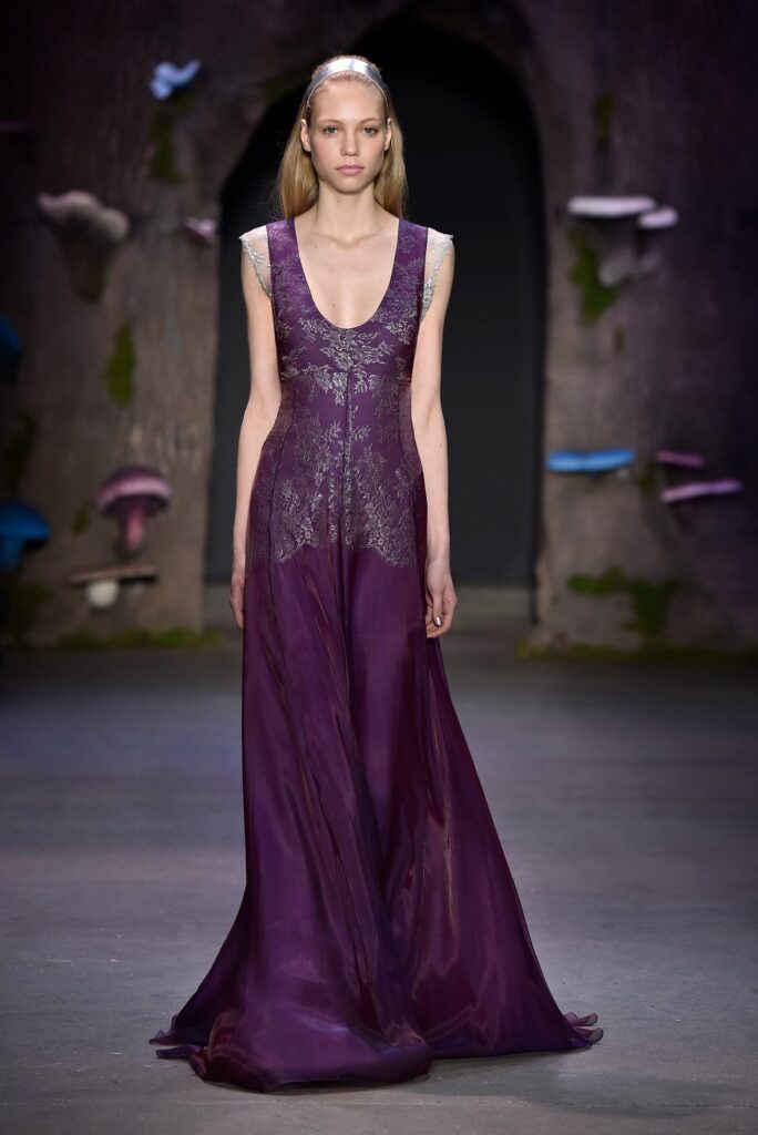 Model in a long, sleeveless purple gown with lace details, walking on a runway with a forest-themed backdrop.