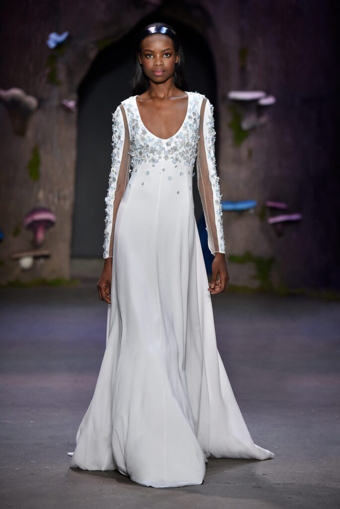 Model in a flowing white gown with sheer beaded sleeves walks a runway with a dark, whimsical background.
