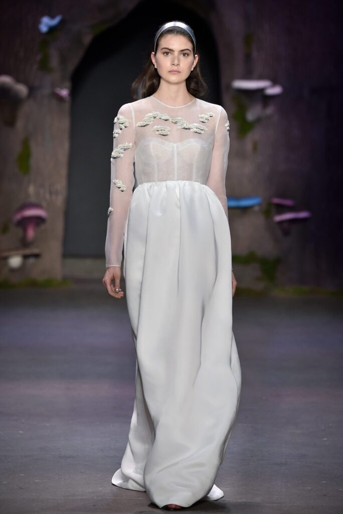 Model in a sheer long-sleeve white gown with applique details, walking on a runway with a dark, whimsical backdrop.