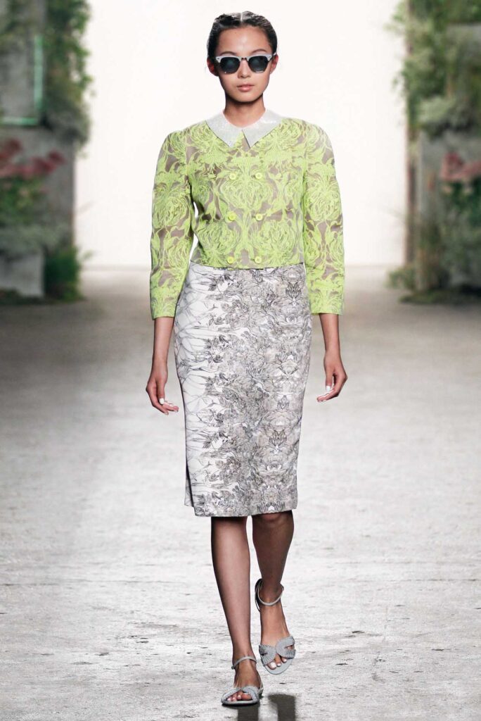 Woman in sunglasses wearing a lime green lace top, floral pencil skirt, and sandals walks on a runway.