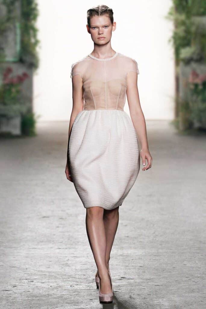Model in a sheer blush top and textured white skirt walks on a runway with floral background.