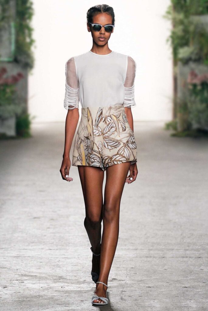 Model in sunglasses wears a white top and patterned shorts, walking on a runway with a foliage backdrop.