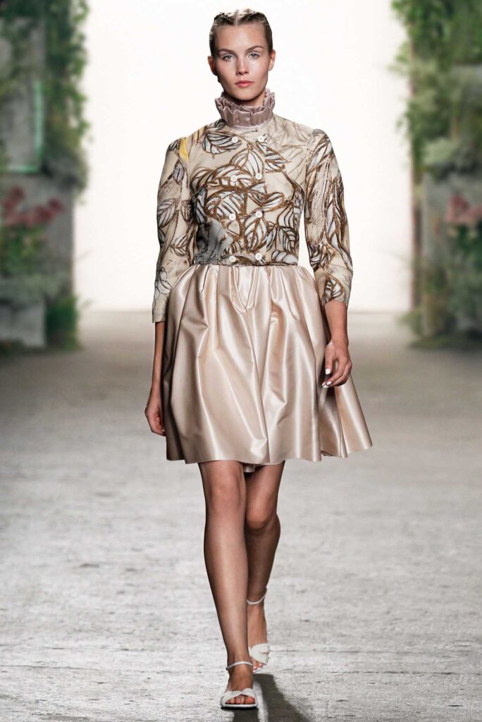 Model in a beige satin dress with floral embroidery walks down a runway with greenery in the background.