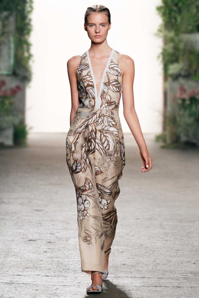 Model wearing a sleeveless, deep V-neck dress with a brown and white floral pattern walks on a runway.