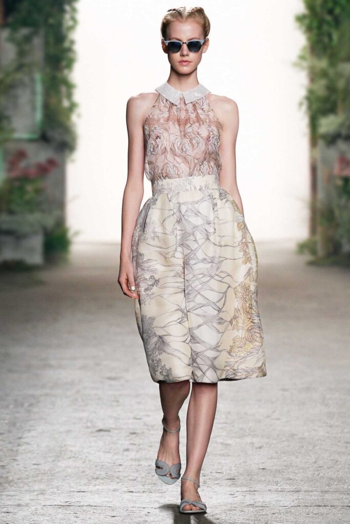 Woman in sleeveless sheer top, full skirt with pattern, sunglasses, and sandals walks a runway.