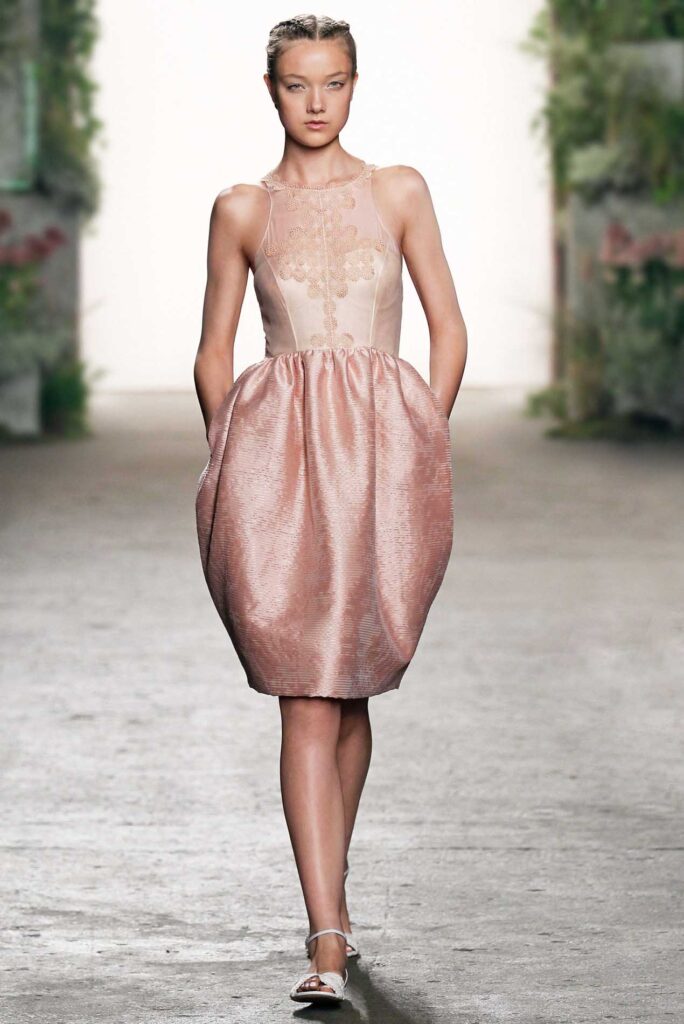 Model walks runway in a sleeveless, blush pink dress with textured skirt, hands in pockets, flat sandals.