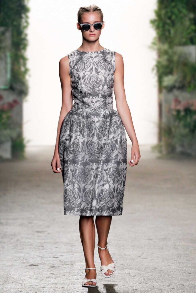 Model in a sleeveless, knee-length, gray floral dress, sandals, and sunglasses walking a runway with greenery.