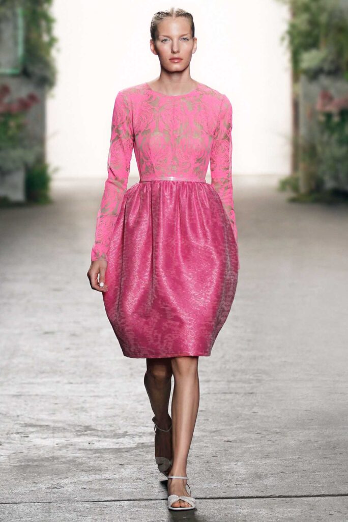 Model walks runway in a bright pink, long-sleeve dress with a patterned bodice and full skirt.