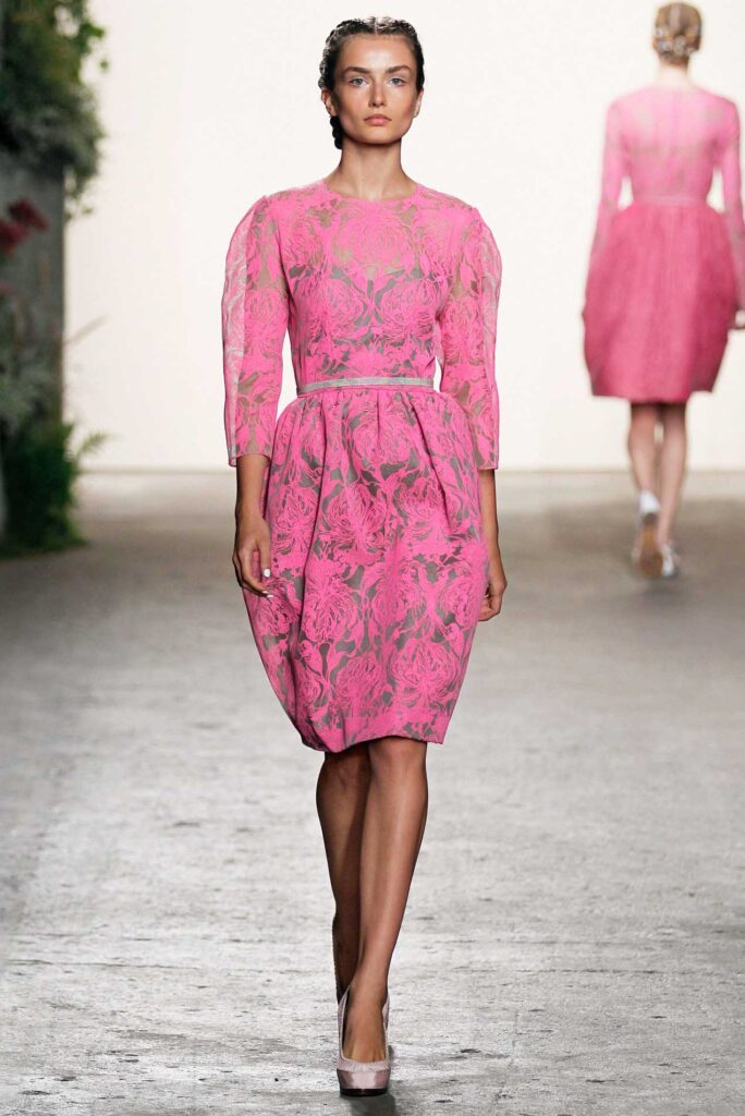 Model walks runway in a pink floral knee-length dress with three-quarter sleeves and matching heels.