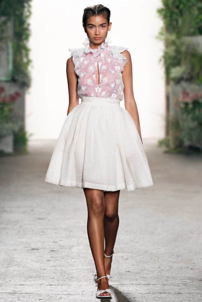 Model in a pink lace top with ruffles and a white flared skirt walks on a runway with greenery in the background.