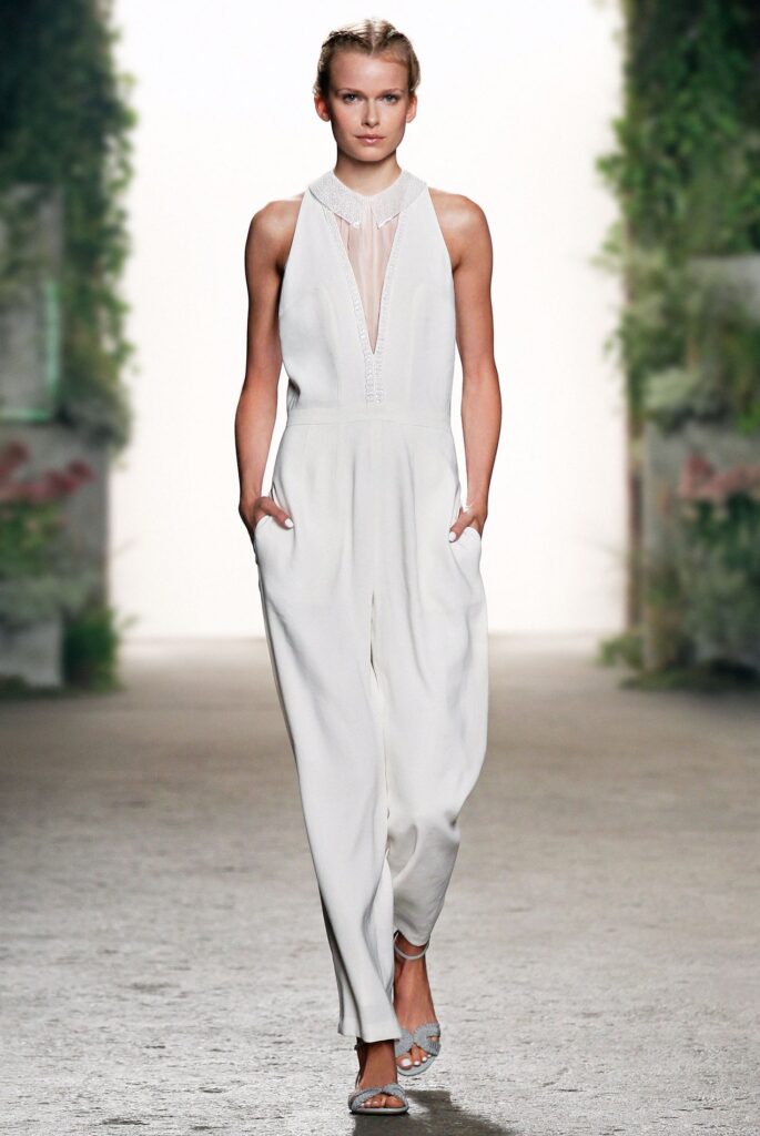 Woman in a sleeveless white jumpsuit with hands in pockets, walking on a runway with a blurred green background.