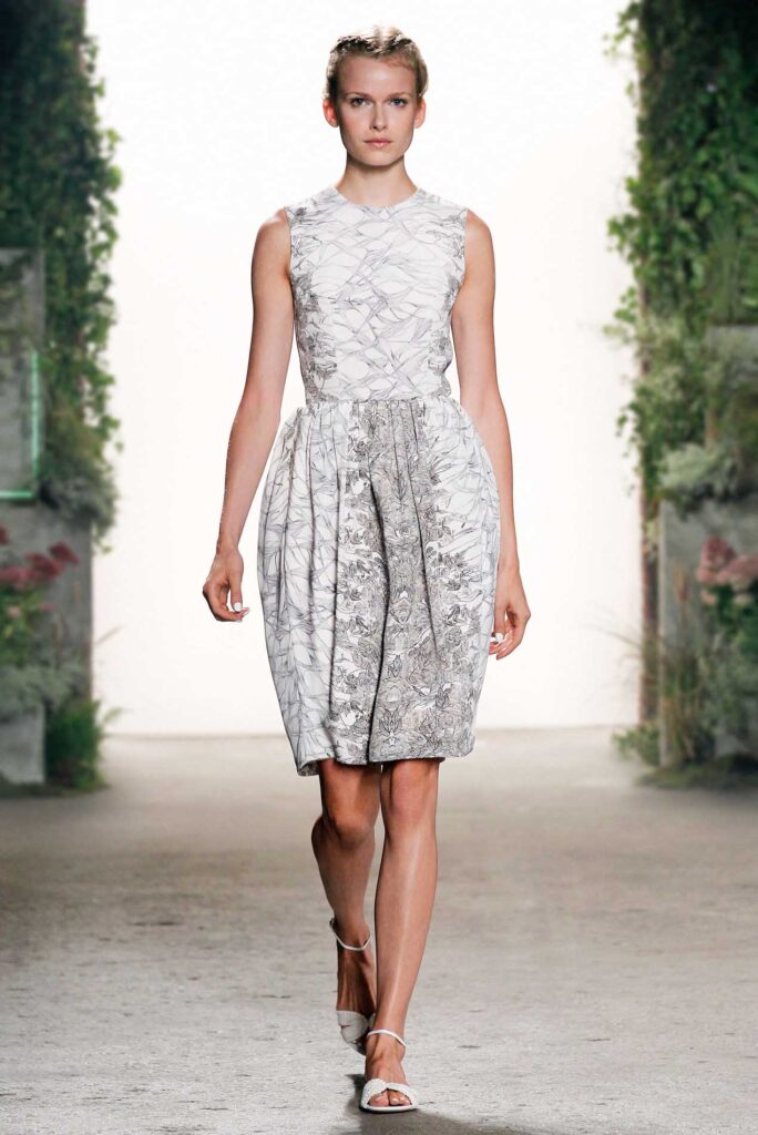 Model walks a runway in a sleeveless, knee-length white dress with a floral pattern and light sandals.