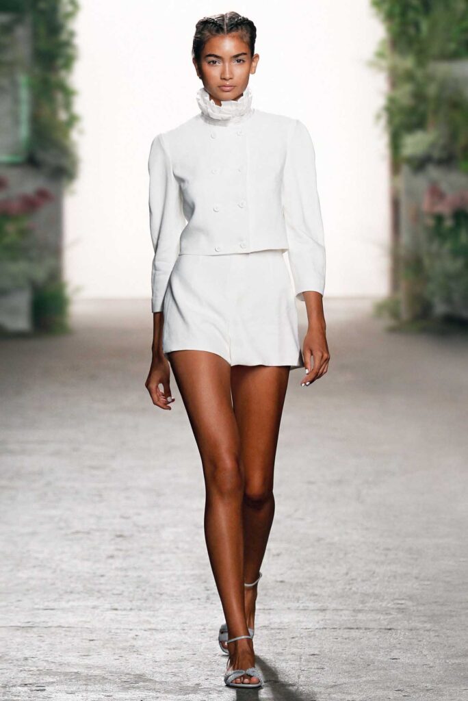 Model in a white, high-neck jacket and shorts walks a runway with greenery in the background.