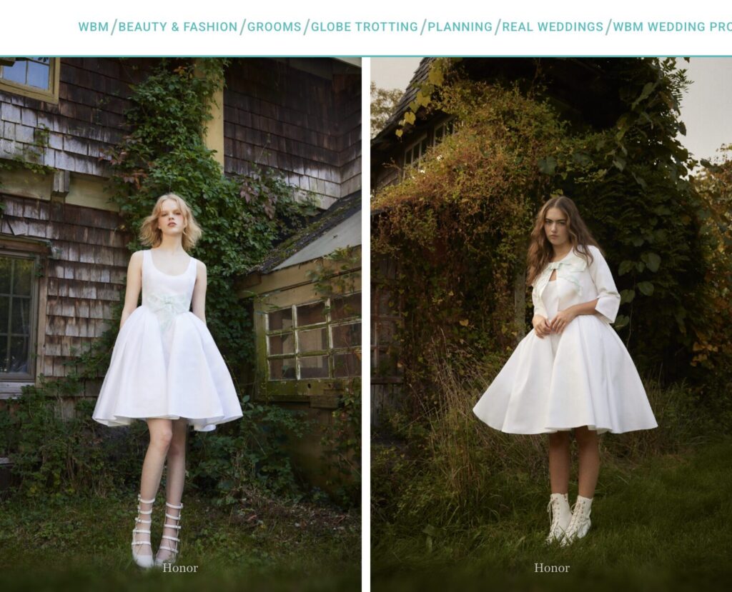Two women in short white bridal dresses pose outdoors near rustic wooden buildings, surrounded by greenery.