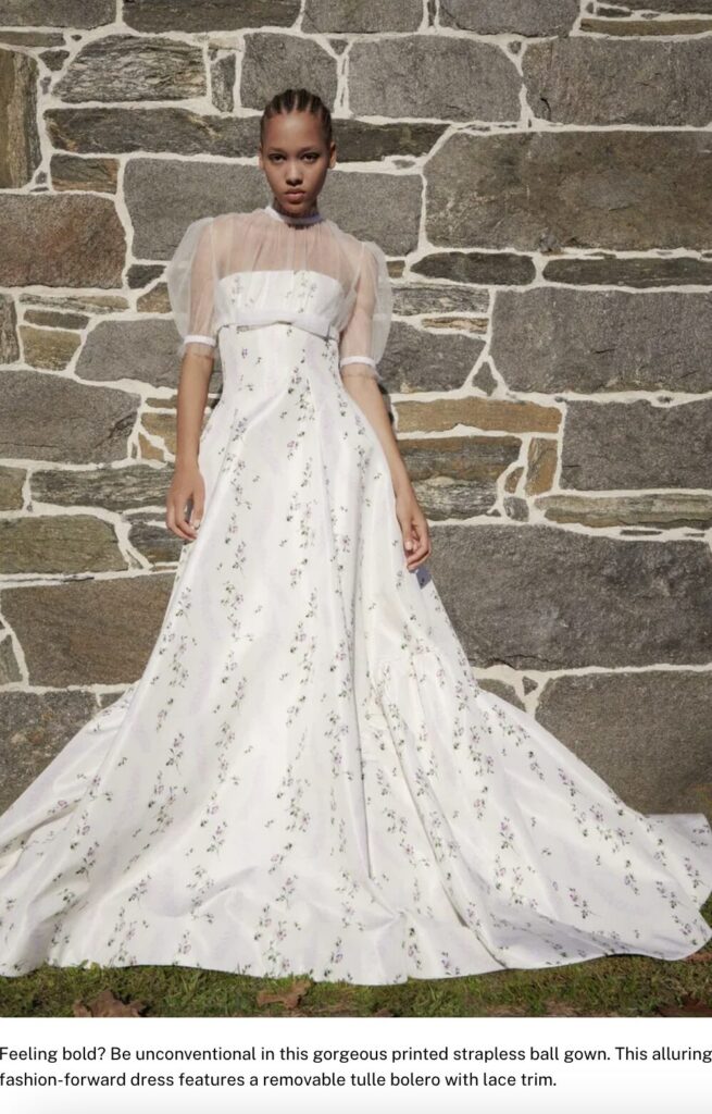 Woman posing in front of a stone wall, wearing a white floral ball gown with sheer sleeves and lace trim.