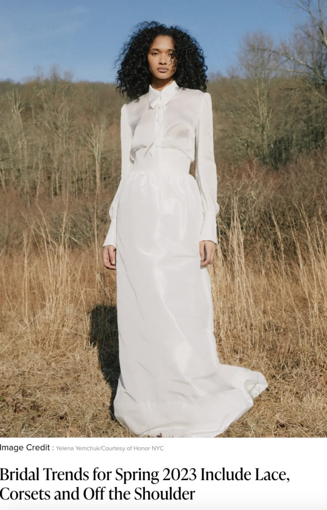 Woman in a long-sleeve white blouse and skirt stands outdoors in a field with dry grass and trees.