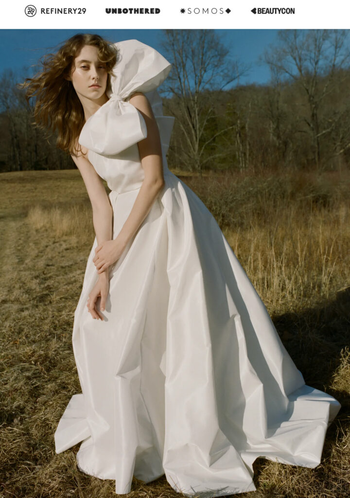 Woman in a white gown stands in a grassy field with trees and blue sky in the background.