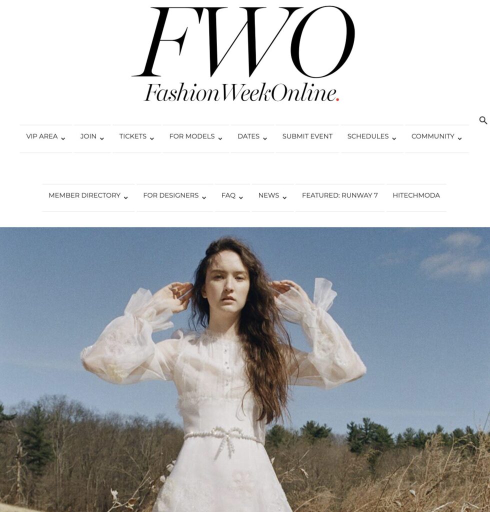 Woman in a white dress stands outdoors under a blue sky, with trees in the background. FashionWeekOnline header above.