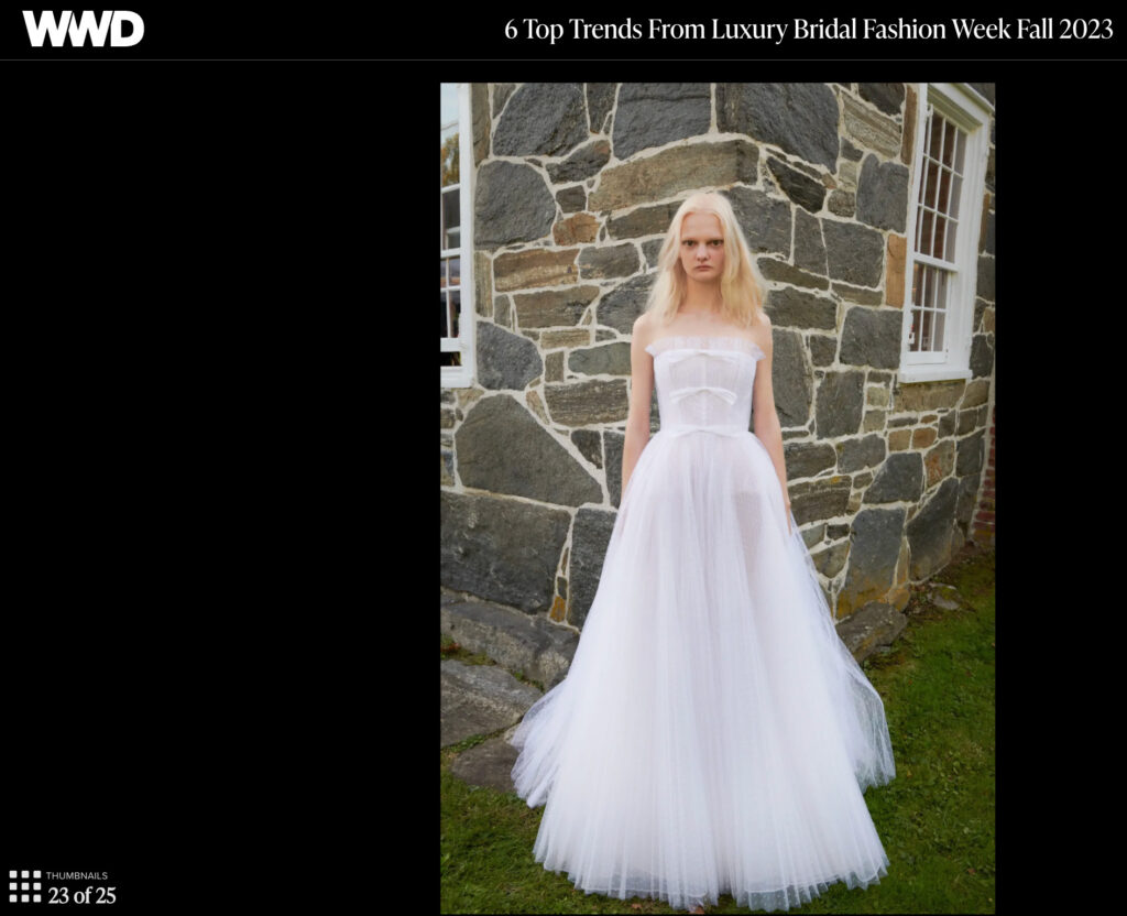 Model in a strapless sheer white wedding gown stands outside by a stone wall on grass.