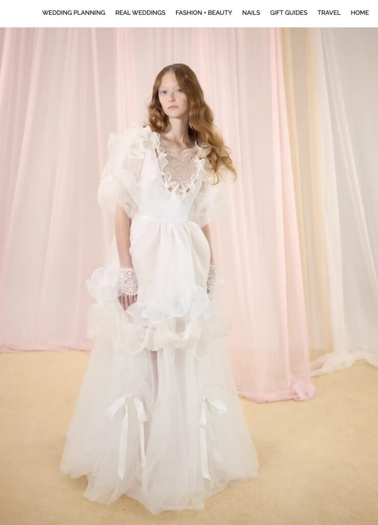 Woman in a ruffled white gown stands on a beige floor with soft pink and white curtains behind her.
