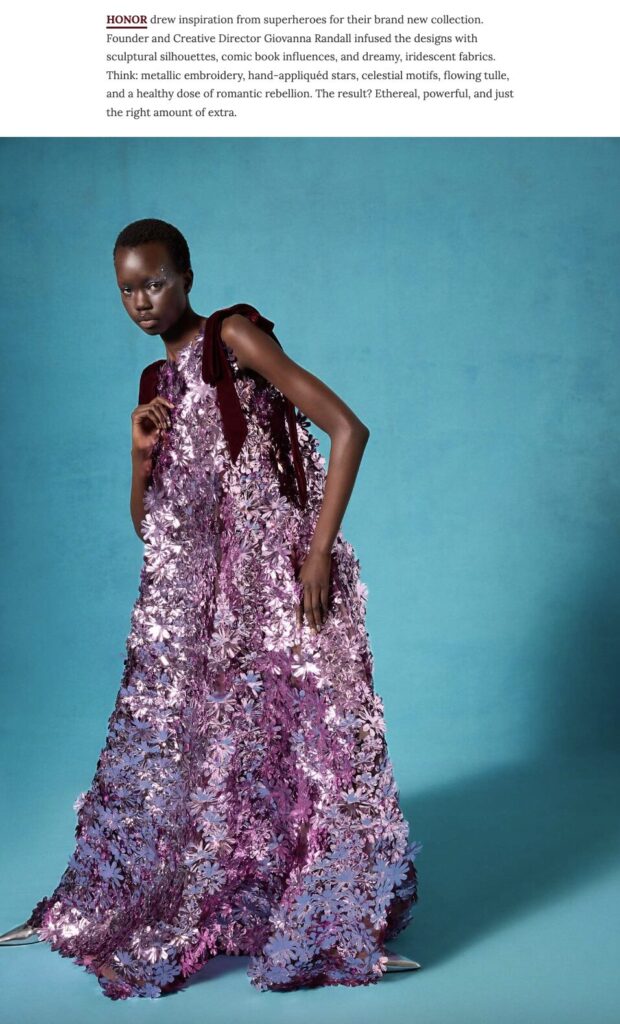 Model poses in a floor-length, purple floral gown with metallic embellishments against a blue backdrop.