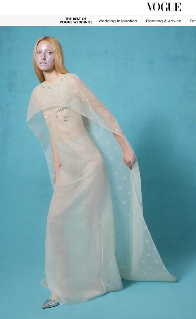 A woman in a sheer, sparkly white gown with a cape poses against a blue background.