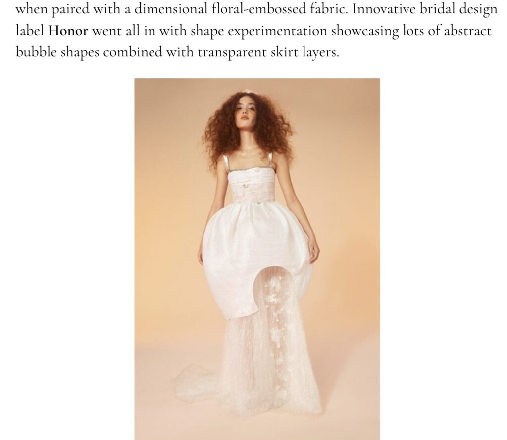 Model in a white strapless gown with a large bubble-shaped skirt and sheer flowing layers, standing against a beige background.