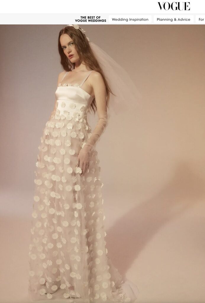 Woman in a white floral wedding gown and veil stands against a soft, neutral background.