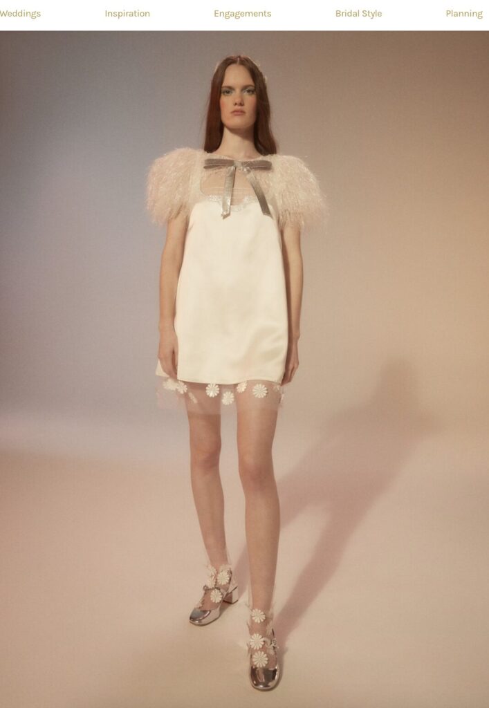 Woman in a short white dress with sheer puff sleeves and floral sandals stands against a neutral background.
