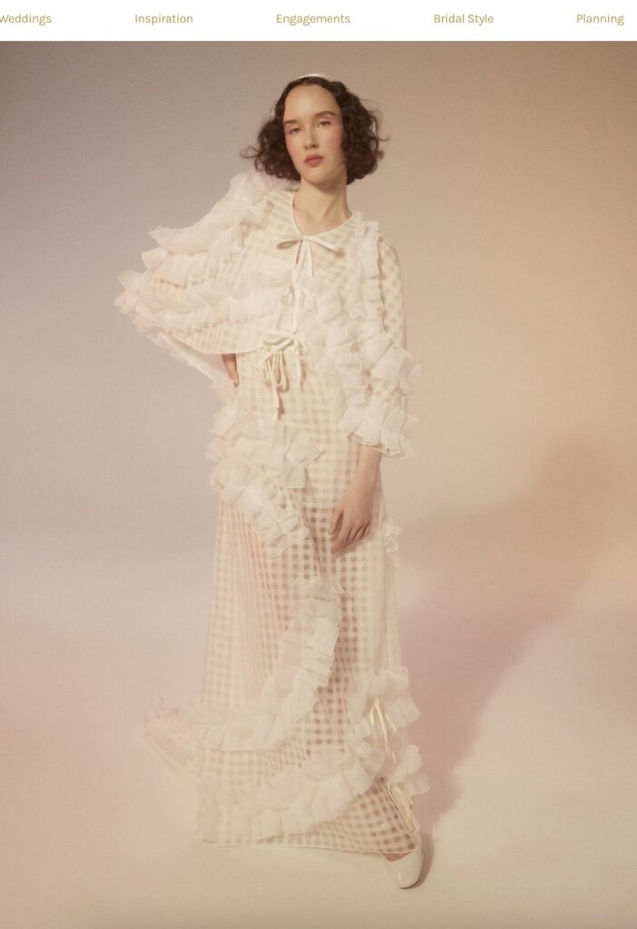 Woman in a sheer, ruffled white dress stands in soft lighting, with a gentle, neutral background.