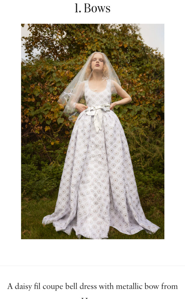 Woman in a white floral gown with a bow and veil stands outdoors in front of green bushes.